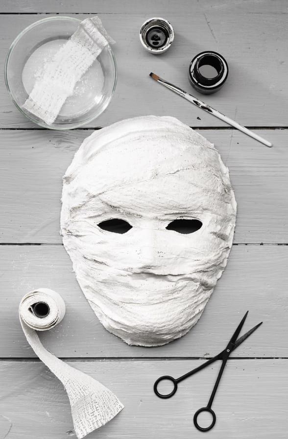 Máscara de gesso em cima de uma mesa clara, ao lado de gaze, tigela de água e tesoura